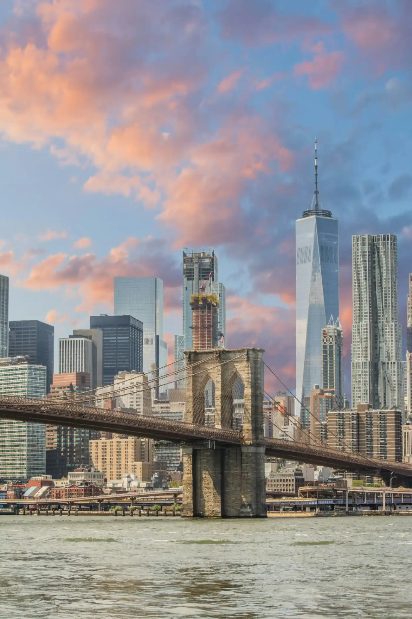 The Brooklyn Bridge with One World Trade Center and the Lower Manhattan skyline under a pink-and-blue sunset sky, representing the New York residents Sanctuary Tulum serves at our oceanfront licensed luxury wellness center in Tulum